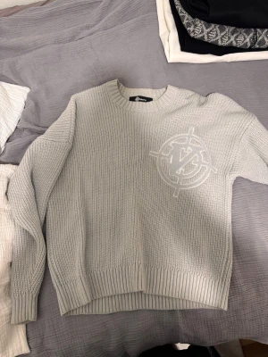Ljusgrå stickad tröja från Vicinity - Snygg ljusgrå stickad tröja från Vicinity med ett stort broderat logomärke på bröstet. Tröjan har rund halsringning, ribbade muddar och är tillverkad i ett grovstickat material som ger en modern och avslappnad look.