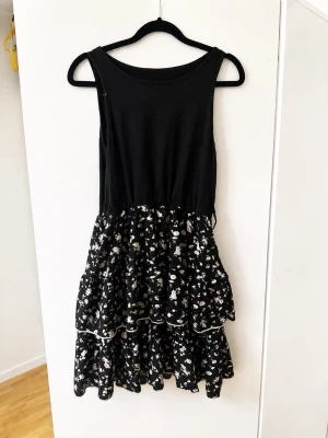 Svart blommig kortklänning - Snygg svart ärmlös klänning med volangig kjol i blommigt mönster. Överdelen är enkel och enfärgad medan kjolen har vita små blommor och flera lager. Perfekt för dig som gillar en lekfull och trendig stil.