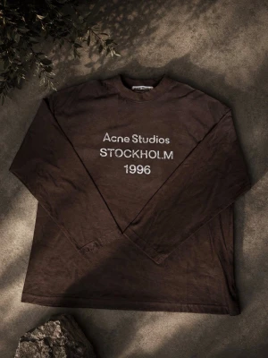 Acne studios long sleeve - Säljer en brun långärmad topp från Acne Studios med tryck 'STOCKHOLM 1996' på bröstet. Klassisk rund halsringning och relaxed fit. Perfekt för dig som gillar minimalistisk och stilren design.