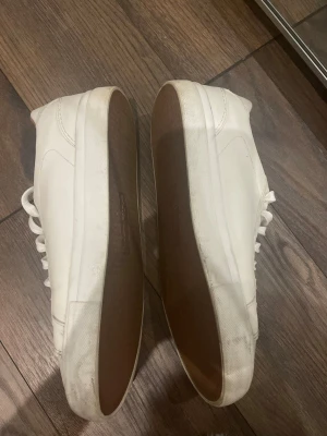 Vita klassiska låga sneakers - Säljer ett par stilrena vita sneakers med lågt skaft och vita snören. Skorna har en slät ovandel i läder och en vit sula med brunt greppmönster undertill. Insidan är ljusbrun och designen är minimalistisk, perfekt för en clean look.