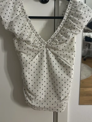 Vit prickig blus med volang - Supersöt vit blus med svarta prickar och volangdetaljer vid axlarna. Djup v-ringning och rynkade sömmar framtill ger en feminin look. Andvänd några få gånger