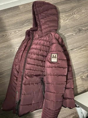 Vinröd dunjacka med huva Marikoo - Snygg vinröd dunjacka från Marikoo med huva och mjukt foder i fuskpäls. Jackan har dragkedja framtill, två fickor med dragkedja och en patch på ärmen. Storlek S.