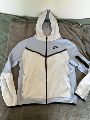 Nike hoodie i vitt och ljusblått - Nike hoodie med dragkedja i vitt och ljusblått. Snygg svart detaljlinje över bröstet och svart Nike-logga på vänster sida. Huva och fickor framtill samt en extra ficka med dragkedja på ena ärmen. Tillverkad i mjukt och bekvämt material.