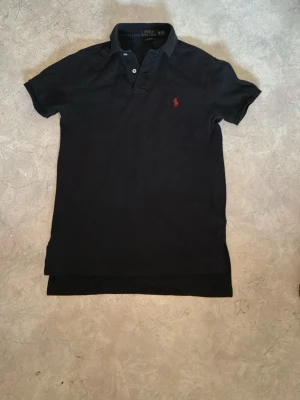 Svart pikétröja från Polo Ralph Lauren - Svart pikétröja i bomull från Polo Ralph Lauren med korta ärmar, klassisk krage och knappar vid halsen. Tröjan har en röd broderad logga på bröstet och en normal passform som ger en clean och tidlös stil. Säljer för jag har en grå istället som jag använder så dennna ligger ba i garderoben sen jag köpte den den är xs men känns lite mer som s i längden då priset kan diskuteras👍
