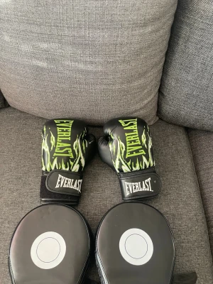 Boxningshandskar och mittsar Everlast - Säljer ett set med boxningshandskar och mittsar från Everlast. Handskarna är svarta med gröna flames och vit logga, tillverkade i konstläder. Mittsarna är svarta med vit cirkel och justerbara band på baksidan. Perfekt för träning och sparring.