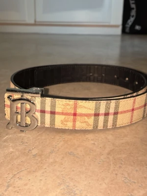 Beige och rutig bälte - Snyggt bälte i beige med klassiskt rutigt mönster i rött och svart. Bältet har ett stort metallspänne framtill. Perfekt accessoar för att lyfta din outfit. Säljer för använde bara mina andra bälten inte denna så har mest luggit i garderoben priset kan diskuteras👍