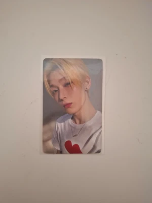 K-pop photocard Romance: Untold - Säljer en K-pop photocard från serien Romance: Untold. Kortet har en bild på en blond idol med vit t-shirt och rött hjärta, samt text på baksidan. Perfekt för samlare eller fans av K-pop.