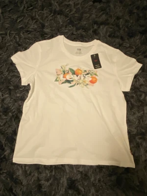 Vit Levi's t-shirt med fruktmotiv - Snygg vit t-shirt från Levi's med färgglatt tryck av apelsiner och blommor på bröstet. Klassisk passform och rund halsringning. Perfekt till jeans eller shorts för en fräsch look.passar även M