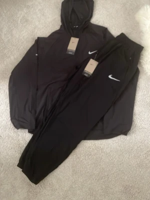 Nike Miller  - Snyggt svart set från Nike med matchande hoodie och byxor. Båda plaggen har den klassiska vita Nike-loggan på bröstet och benet. Setet är tillverkat i mjukt material och har en avslappnad passform. Perfekt för en sportig och chill stil.