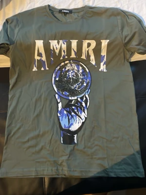 Grön AMIRI t-shirt med tryck - Snygg grön t-shirt från AMIRI med stort grafiskt tryck framtill som visar en hand som håller en klot och AMIRI-loggan i vitt och blått ovanför. Klassisk rund hals och korta ärmar. Perfekt för dig som vill sticka ut med en unik design.