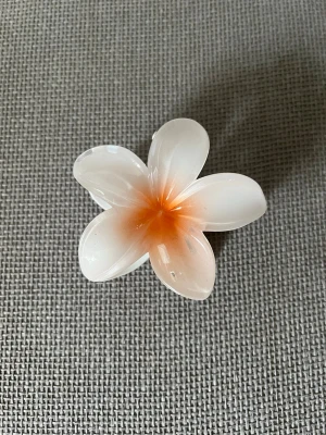 Blomformad hårklämma i plast - Söt hårklämma i form av en blomma med fem kronblad. Klämman är vit med en mjuk orange ton i mitten och har en glansig finish. Tillverkad i plast och perfekt för att ge en somrig touch till din frisyr.