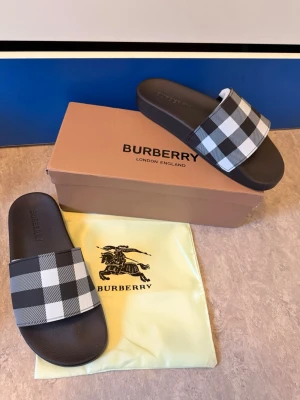 Svarta tofflor med rutigt mönster Burberry - Snygga svarta tofflor från Burberry med klassiskt svartvitt rutigt mönster på remmen. Tillverkade i syntetmaterial med präglad Burberry-logga på fotbädden. Perfekta för en avslappnad och trendig stil.