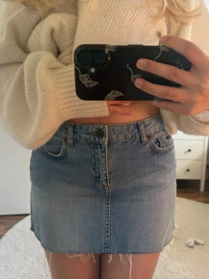 Ljusblå jeanskjol med rå kant - Trendig ljusblå jeanskjol i kort modell med rå, fransig kant nedtill. Kjolen har klassisk femficksdesign, bälteshällor och knappgylf. Perfekt för en avslappnad och cool stil.