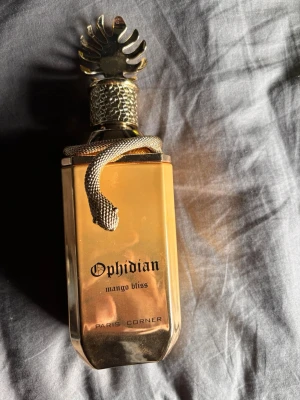 Ophidian Mango Bliss parfymflaska - Lyxig parfymflaska i guld med unikt ormformat lock och detaljer. Flaskan har en elegant, fyrkantig form och texten 'Ophidian mango bliss' på framsidan. Perfekt för dig som gillar exklusiva och iögonfallande dofter.