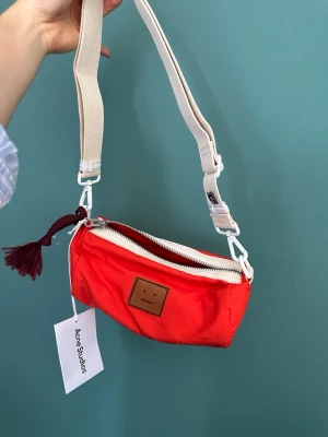 Röd axelväska från Acne Studios - Cool röd axelväska från Acne Studios i nylon med justerbar beige axelrem och vit dragkedja. Väskan har Acne Studios klassiska smiley-logga framtill och en dekorativ tofs vid dragkedjan. Perfekt för att bära det viktigaste med stil.