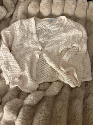 Beige stickad kofta från Isolde - Söt beige stickad kofta från Isolde i storlek M. Koftan har ett fint hålmönster, lång ärm och en enkel knapp framtill. Perfekt att slänga över en topp för en chill look. Mjukt material och avslappnad passform.