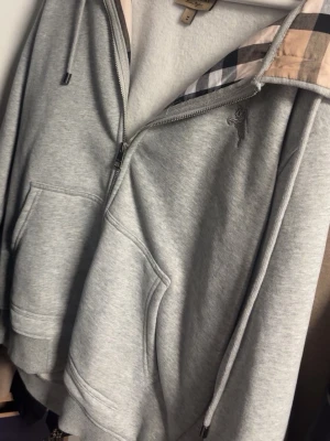 Grå hoodie från Burberry med huva - Snygg grå hoodie från Burberry med dragkedja, klassiskt rutigt foder i huvan och två stora fickor framtill. Tillverkad i mjukt sweatshirtmaterial och har justerbar dragsko i huvan. Perfekt för en chill och stilren look.