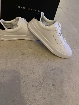 Vita sneakers från Tommy Hilfiger - Stilrena vita sneakers från Tommy Hilfiger med lågt skaft och svarta detaljer vid hälen. Skorna har klassisk snörning och är tillverkade i slätt lädermaterial. Diskret logga på sidan ger en clean look som passar till det mesta.