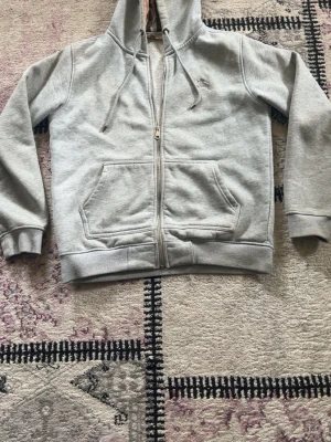 Grå zip hoodie från Burberry - Säljer en grå hoodie från Burberry med dragkedja, huva och klassiskt rutigt foder i luvan. Hoodien har två stora fickor framtill och broderad logga på bröstet. Mjuk insida i fleece och ribbade muddar vid ärmar och nederkant.