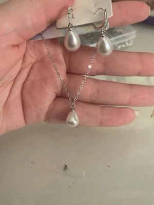Set med silverfärgade pärldroppar - Säljer ett matchande set med halsband och örhängen i silverfärg. Både halsbandet och örhängena har droppformade pärlor som ger en elegant och stilren look. Perfekt för dig som gillar klassiska accessoarer med en modern twist.