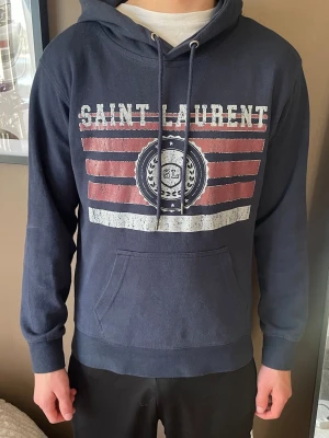 Mörkblå Saint Laurent hoodie och dondup jeans - Mörkblå hoodie från Saint Laurent med tryck i vitt och rött framtill. Klassisk huva med dragsko och stor magficka. Mjuk bomullskänsla och ribbade muddar vid ärmslut och nederkant. Perfekt för en avslappnad och trendig look.