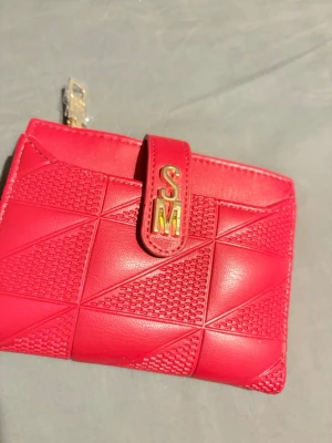Röd plånbok från Steve Madden - Cool röd plånbok från Steve Madden med guldfärgade SM-detaljer. Plånboken har ett geometriskt mönster, flera kortfack och ett fönster för ID. Tillverkad i konstläder och stängs med tryckknapp.