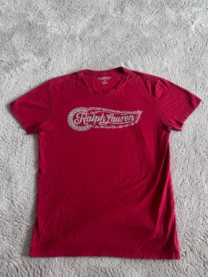 Röd t-shirt från Ralph Lauren - Säljer en röd t-shirt från Ralph Lauren med tryckt logga i grått framtill. T-shirten har rund halsringning och korta ärmar. Tillverkad i mjuk bomull, perfekt för en avslappnad stil.
