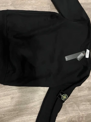 Svart sweatshirt från Stone Island - Säljer en svart sweatshirt i storlek S från Stone Island med den klassiska logotypen på ärmen. Tröjan har rund halsringning, långa ärmar och ribbade muddar. Perfekt för dig som gillar streetwear och vill ha en stilren look.