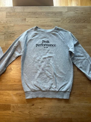 Grå sweatshirt från Peak Performance - Klassisk grå sweatshirt från Peak Performance med svart logga och text på bröstet. Tröjan har rund hals, långa ärmar och ribbade muddar vid ärmslut och nederkant. Skön tröja som inte används så mycket. Den är i barn storleken 140. 