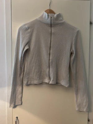 Ljusgrå ribbad ziptröja med hög krage -  ljusgrå ribbad tröja med dragkedja framtill och hög krage. Långärmad och croppad modell som passar perfekt till jeans eller kjol. Materialet är mjukt och stretchigt för en bekväm känsla.