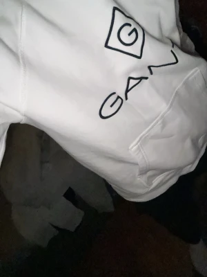 Vit hoodie från GANT med logga - Säljer en stilren vit hoodie från GANT med stor svart logga och grafiskt tryck på bröstet. Hoodien har huva och en klassisk magficka. Perfekt för dig som gillar enkel men snygg streetwear. Materialet känns mjukt och skönt mot huden.