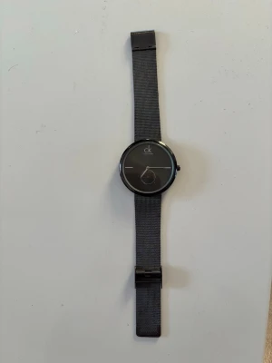 Svart klocka från Calvin Klein - Stilren svart klocka från Calvin Klein med minimalistisk urtavla och mesh-armband i metall. Klockan har en rund form och diskret logga på urtavlan. Perfekt accessoar för en modern look. Behöver nytt batteri 