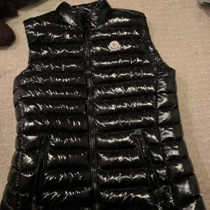 Svart glansig dunväst från Moncler - Snygg svart dunväst från Moncler med glansig finish och klassisk logga på bröstet. Västen har två dragkedjefickor framtill och hög krage. Perfekt att slänga över en hoodie eller tröja för en cool look. Nästan aldrig använd, har dock inget kvitto kvar men den är äkta.