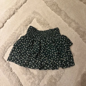 Blommig volangkjol från Zara - Supersöt kort kjol från Zara med blommigt mönster i vitt på mörkgrön botten. Kjolen har volanger och resår i midjan för en bekväm passform. Perfekt för dig som gillar en lekfull och trendig stil.