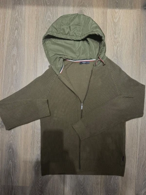 Cardigan från Hechter Paris - Snygg olivgrön stickad cardiganjacka från Hechter Paris med dragkedja och luva. Jackan har ribbstickade muddar och en avslappnad passform. Tweka inte, fråga gärna och kom med pris förslag så löser vi något tillsammans!