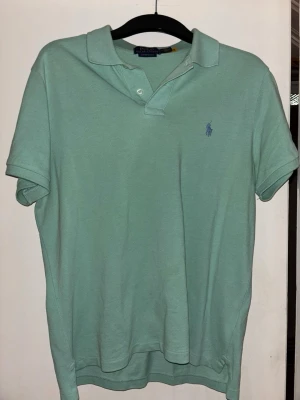 Mintgrön pikétröja från Ralph Lauren - Fräsch mintgrön pikétröja från Polo Ralph Lauren med klassisk krage och två knappar. Tröjan har broderad logga i blått på bröstet och är tillverkad i mjuk bomull. Perfekt för en stilren och avslappnad look.