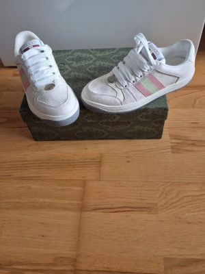 Vita Gucci sneakers med rosa/gröna ränder - Snygga vita sneakers från Gucci med klassiska rosa och gröna ränder på sidan. Skorna har vita snören, metallfärgad GG-detalj framtill och logga på plösen. Tillverkade i canvas och läder för en clean och exklusiv look.