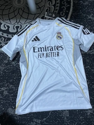 Real Madrid matchtröja Adidas XL - Säljer en vit Real Madrid matchtröja från Adidas i storlek XL. Tröjan har svarta detaljer, gula linjer längs sidorna och klubbmärke på bröstet. Tillverkad i lätt och ventilerande material, perfekt för fotboll eller träning.
