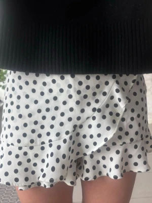 Vit prickiga shorts med volang - Supersöt  vita shorts som ser ut som en kjol med svarta prickar och volangkant. Omlottkänsla framtill och mjukt, lätt tyg som ger en flowy look. Perfekt för dig som gillar lekfulla detaljer och trendiga mönster.