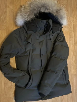 Mörkgrön dunjacka från Canada Goose - Säljer en grå dunjacka från Canada Goose med stor huva och avtagbar pälskant. Jackan har en klassisk patch på ärmen, dragkedja och knappar framtill samt flera fickor. Perfekt för kalla vinterdagar och riktigt snygg streetstil. Säljer pga sällan används.                                                                PRISET ÄR EJ HUGGET I STEN!