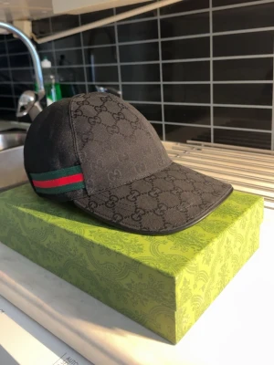 Svart Gucci keps med GG-mönster - Snygg svart keps från Gucci med klassiskt GG-monogram över hela kepsen. På sidan finns den ikoniska gröna och röda randen. Justerbar rem bak i läder och böjd skärm. Tillverkad i canvasmaterial som ger en exklusiv känsla.