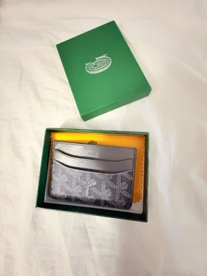 Grå korthållare - Säljer ett stilrent svart kortfodral från G med det klassiska vita mönstret och loggan framtill. Fodralet har flera fack för kort och är tillverkat i exklusivt läder och canvas. Kommer i original grön ask med tillhörande certifikat.
