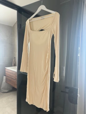 Ljusbeige långärmad klänning från Oh Polly - Trendig ljusbeige klänning med lång ärm och fyrkantig ringning. Klänningen har snörningsdetaljer vid ärmsluten och en öppen rygg med snygg cut-out. Materialet känns mjukt och stretchigt