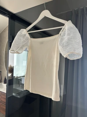 Vit topp med puffärmar - Trendig vit topp med fyrkantig urringning och genomskinliga puffärmar i mesh. Kroppen är i mjukt tyg och toppen har en croppad passform. 