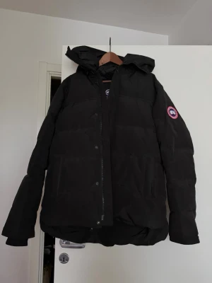 Svart dunjacka från Canada Goose XL - Säljer en svart dunjacka från Canada Goose i storlek XL. Jackan har en stor huva, två stora framfickor och dragkedja med vindslå. Perfekt för kalla vinterdagar och riktigt varm tack vare dunfyllningen. Klassisk och stilren design.
