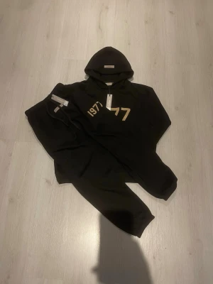 Svart 1977 Essentials hoodie - Jag skickar innan 20:00 och vi kan snacka om priset men Säljer en svart hoodie från Essentials med trycket '1977' i beige framtill. Hoodien har en klassisk huva och känguruficka. Perfekt för dig som gillar streetwear och vill ha en stilren look. Materialet känns mjukt och skönt, och passformen är relaxed.