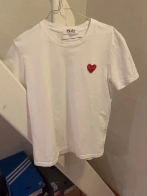 Vit Comme des Garçons PLAY t-shirt. Cdg T-shirt - Vit t-shirt från Comme des Garçons PLAY med det ikoniska röda hjärtat med ögon broderat på bröstet. Klassisk passform och rund halsringning. Perfekt basic-plagg med en twist, tillverkad i mjuk bomull. Tveka inte på att höra av dig om du undrar något!🙂