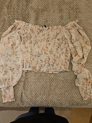 Blommig offshoulder blus Gina Tricot - Supersöt offshoulder blus från Gina Tricot i storlek 36. Blusen är ljus med små blommor i gult, blått och orange, har smockad kropp och volangdetaljer vid ärmslut och nederkant. Långa puffärmar i tunn, luftig kvalitet. Perfekt för en trendig look!