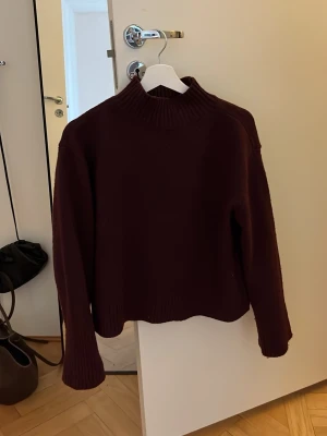 Vinröd stickad tröja från Ellos - Mysig vinröd stickad tröja från Ellos med hög krage och långa ärmar. Tröjan har en rak passform och ribbstickade detaljer vid hals, ärmslut och nederkant. Perfekt för kyliga dagar och enkel att styla till olika outfits.