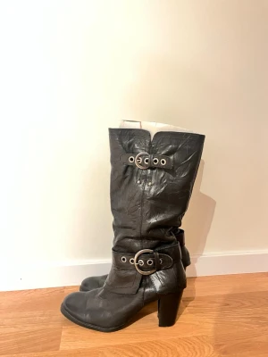 Svarta höga läderstövlar med spännen - Säljer ett par svarta höga stövlar i äkta läder med chunky klack och dekorativa metallspännen både upptill och vid ankeln. Stövlarna har rundad tå och en cool, lite slouchy passform som ger en edgy vibe. Perfekta till jeans eller kjol för en trendig look.